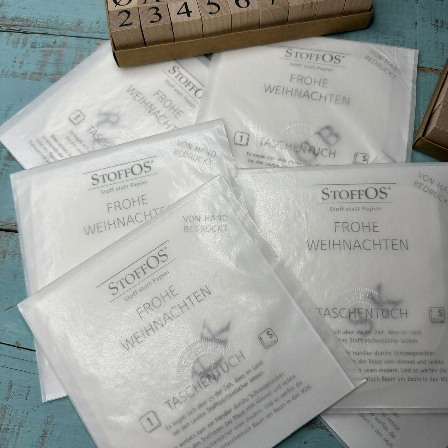 Einzelne weiße Stofftaschentücher von Hand mit Initialen bedruckt, in Pergamintüten verpackt hier mit saisonalem Gruß auf der Verpackung: Frohe Weihnachten.