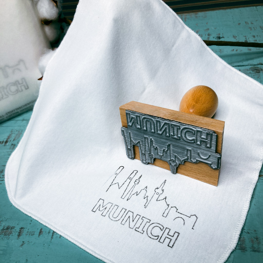 Tolles Souvenir für München Fans. Auf dem StoffOS Taschentuch ist die stilisierte Shilouette der Sehenswürdigkeiten von München unten rechts in schwarz grau aufgedruckt, Darüber liegt der Stempel als Anschauungsobjekt.