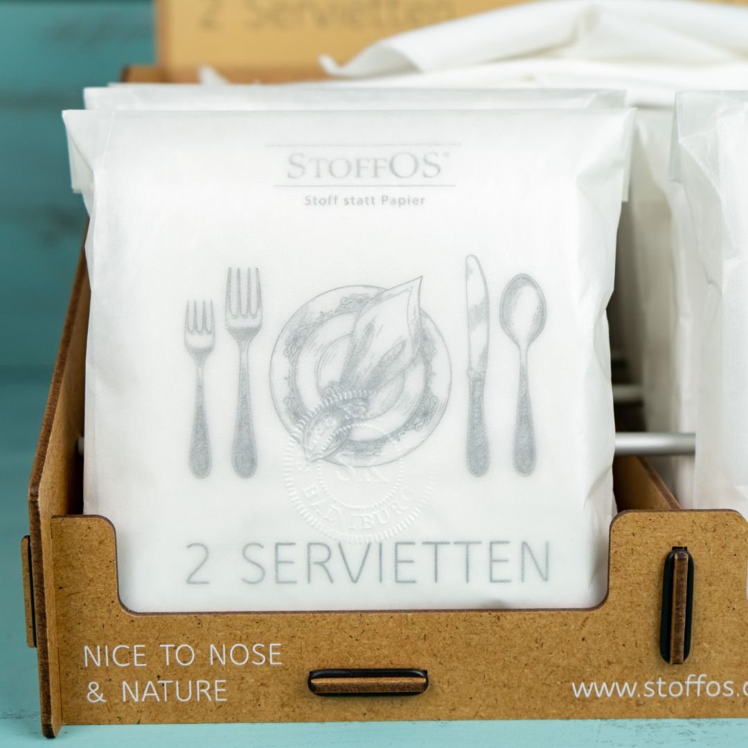 Stoffservietten verpackt zeigt hier die Frontseite der 2er Packs der Lippenschmeichler. Die Pergamintüte steht in einem Verkaufsdisplay. Auf der Verpackung steht unter dem StoffOS Firmenlogo der Slogan: Stoff statt Papier. Darunter ist eine historisch anmutende Zeichnung eines Gedeckes, ca. 8 cm breit und 6 cm hoch. Links zwei Gabeln, rechts Messer und Löffel. In der Mitte eine Serviette, die mit einem Serviettenring gefasst diagonal auf einem Platzteller liegt.