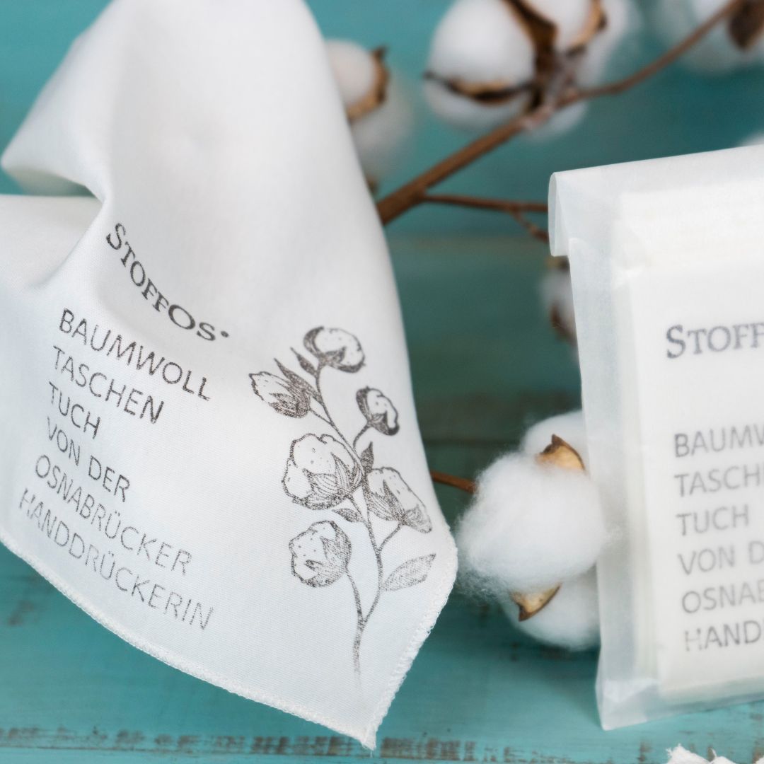 Stofftaschentücher HANDBEDRUCKT 5er Pack Biobaumwolljersey. In Kieselgrau handbedrucktes Stofftaschentuch, genannt StoffO hängt über Baumwollzweigen. Links neben einem 9 cm hohen Baumwollzweig mit Fruchtständen ist der Spruch: „Baumwolltaschentuch von der Osnabrücker Handdrückerin“ in 8 mm hohen Großbuchstaben in sechs Zeilen aufgedruckt Daneben steht eine Pergamintüte mit fünf StoffOS.