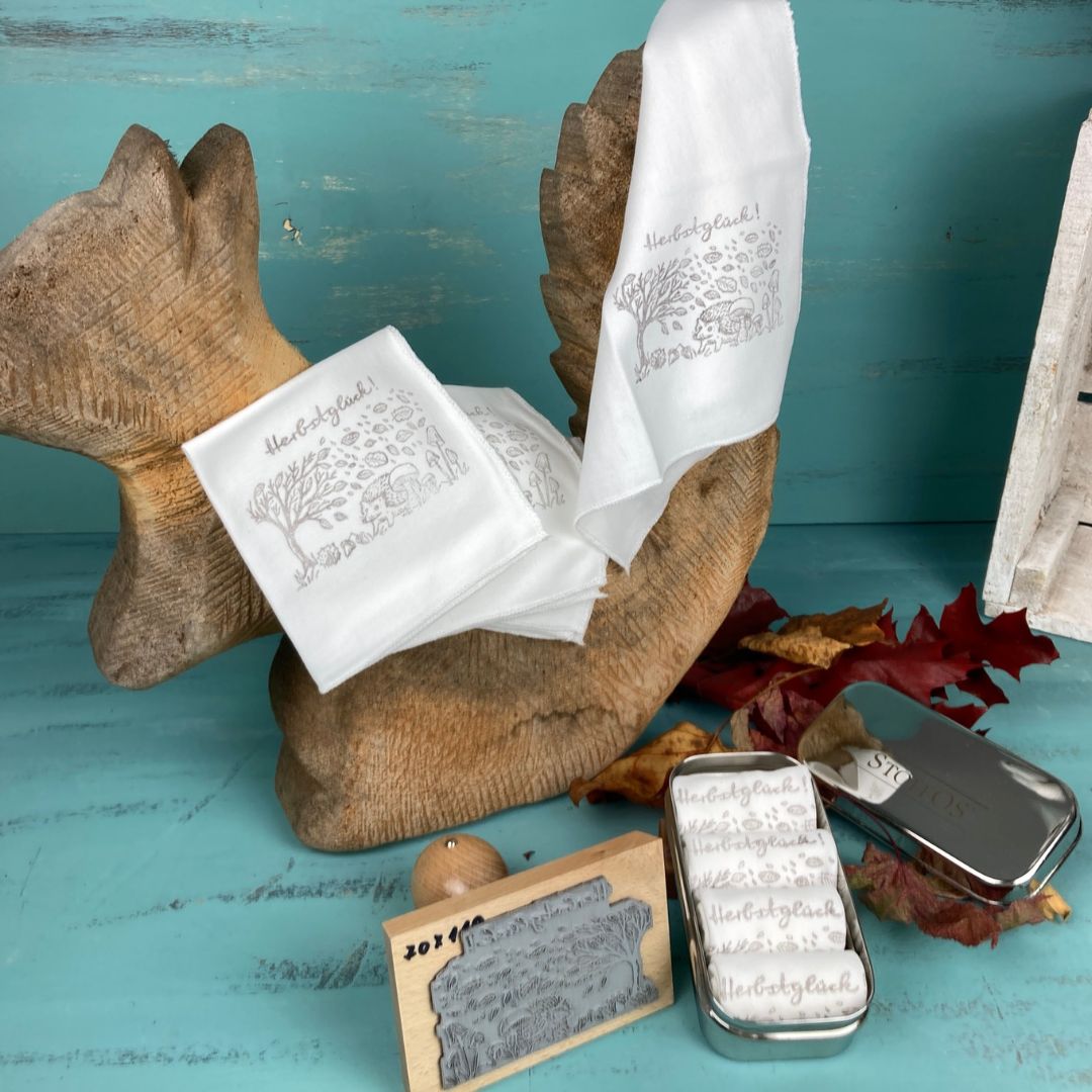 Stofftaschentücher HANDBEDRUCKT 4er Set 22,5 x 22,5 cm Motiv Herbstglück, BioBaumwolle. Ein ausgebreitetes Tuch hängt über einem Eichhörnchen aus Holz, so dass unten rechts das aufgedruckte dunkelgraue Motiv 9 cm breit, 6 cm hoch, der Herbstlandschaft zu sehen ist. Im Vordergrund liegt eine geöffnete Tiny Box mit vier StoffOS Röllchen zwischen dem Stempel und dem Deckel der Box.