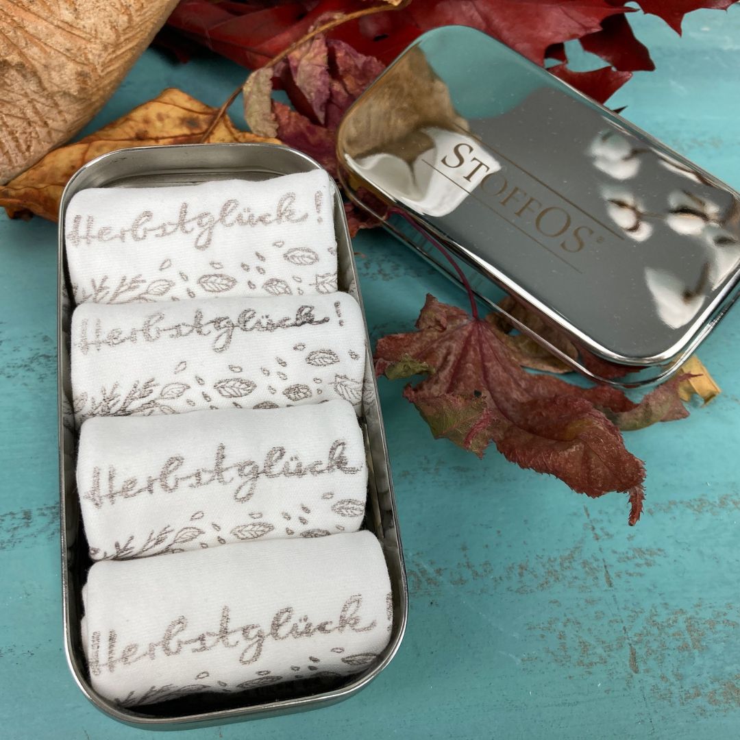 Stofftaschentücher HANDBEDRUCKT aus Biobaumwolljersey. Vier aufgerollte StoffOS mit dem Herbstglück Motiv bedruckt in kieselgrau, liegen nebeneinander in der geöffneten Tiny BOX. Daneben liegt der Deckel, mit dem StoffOS Logo. Buntes Herbstlaub liegt im Hintergrund.  Das Bild hat Nele Jamin von mach-werk aus der Igelperspektive gezeichnet. Links biegt sich ein Baum im Sturm, Blätter wehen durch die Luft. Am Boden die üblichen Herbstgefährten: Kastanien, Eicheln, Pilze und ein Igel.