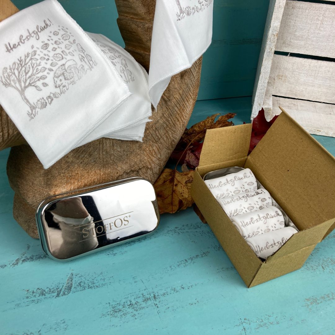 Stofftaschentücher HANDBEDRUCKT 4er Set 22,5 x 22,5 cm Motiv Herbstglück, BioBaumwolle. Ein ausgebreitetes Tuch hängt über einem Eichhörnchen aus Holz, so dass unten rechts das aufgedruckte dunkelgraue Motiv 9 cm breit, 6 cm hoch, der Herbstlandschaft zu sehen ist. Im Vordergrund liegt eine geöffnete Tiny Box mit vier StoffOS Röllchen in der dazugehörigen, geöffneten braunen Pappschachtel. Links liegt der Deckel der Box.