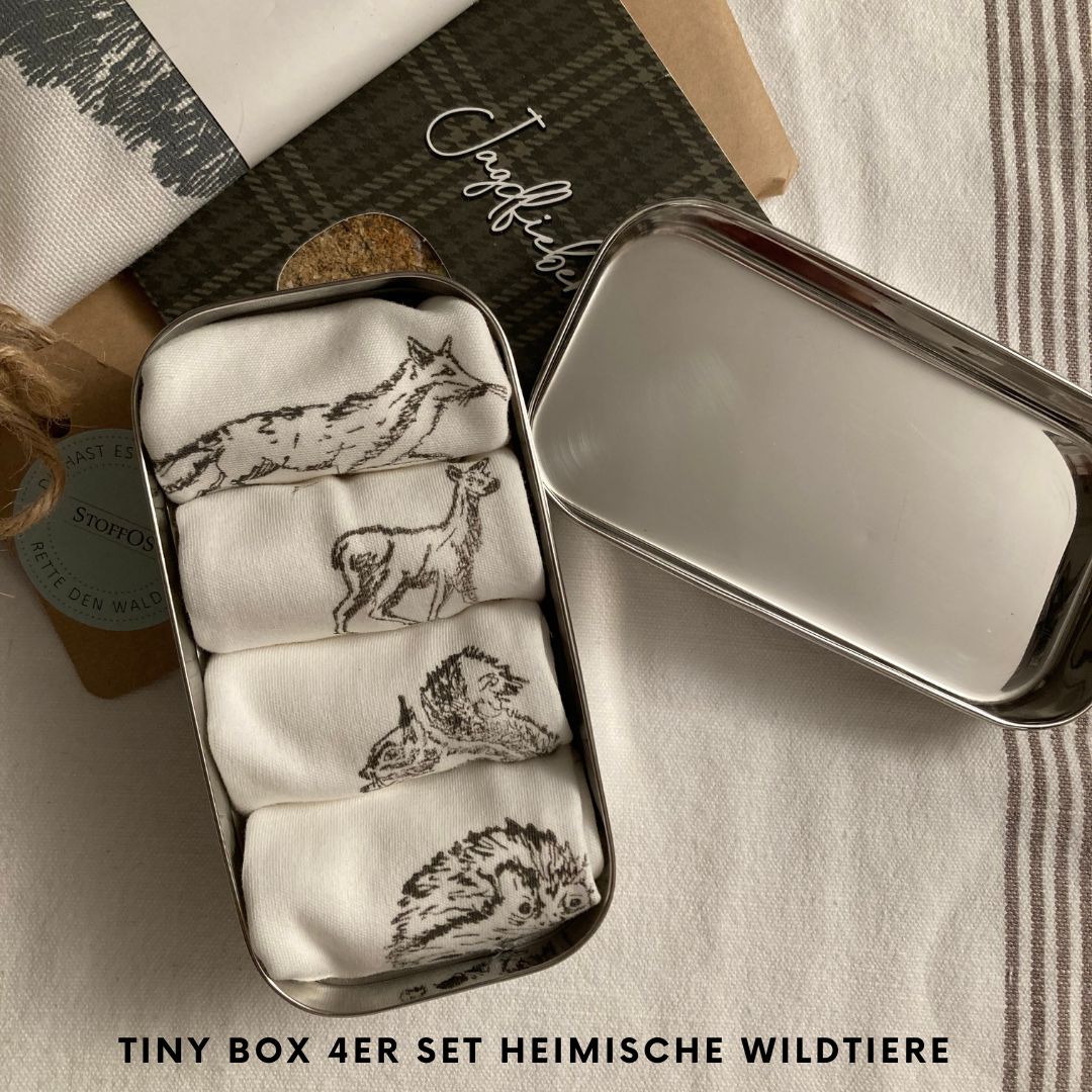 Stofftaschentücher HANDBEDRUCKT. Die geöffnete 6,5 cm breite, 12 cm lange Tiny BOX aus hochglänzendem Edelstahl ist mit Deckel 4 cm hoch. Darin liegen vier aufgerollte Stofftaschentücher, StoffOS genannt nebeneinander. Die einzelnen Rollen sind 6 cm breit. Sie sind einzeln mit jeweils einem Tiermotiv in der Farbe Kieselgrau bedruckt. Die Motive Fuchs, Reh, Eichhörnchen und Igel sind je nach Tierform, 4 x 4 cm groß der Fuchs ist mit 7 cm das breiteste Motiv.