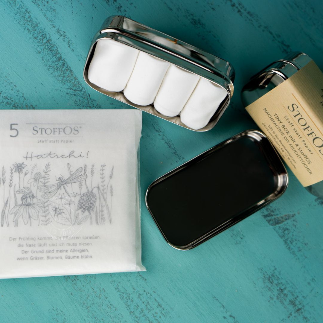 Stofftaschentücher, weiß 4er und 5er Set aus Biobaumwolljersey. Von oben betrachtet. Neben der Hatschi- Pergamintüte mit fünf weißen StoffOS als Inhalt liegt eine geöffnete Tiny Box 12 x 6 x 4 cm groß, mit vier aufgerollten StoffOS. Eine weitere Box, die nicht zum Lieferumfang gehört, ist mit einer Banderole aus Kraftpapier umwickelt. Darauf steht: nachhaltige Do it Yourself Feuchttücher.