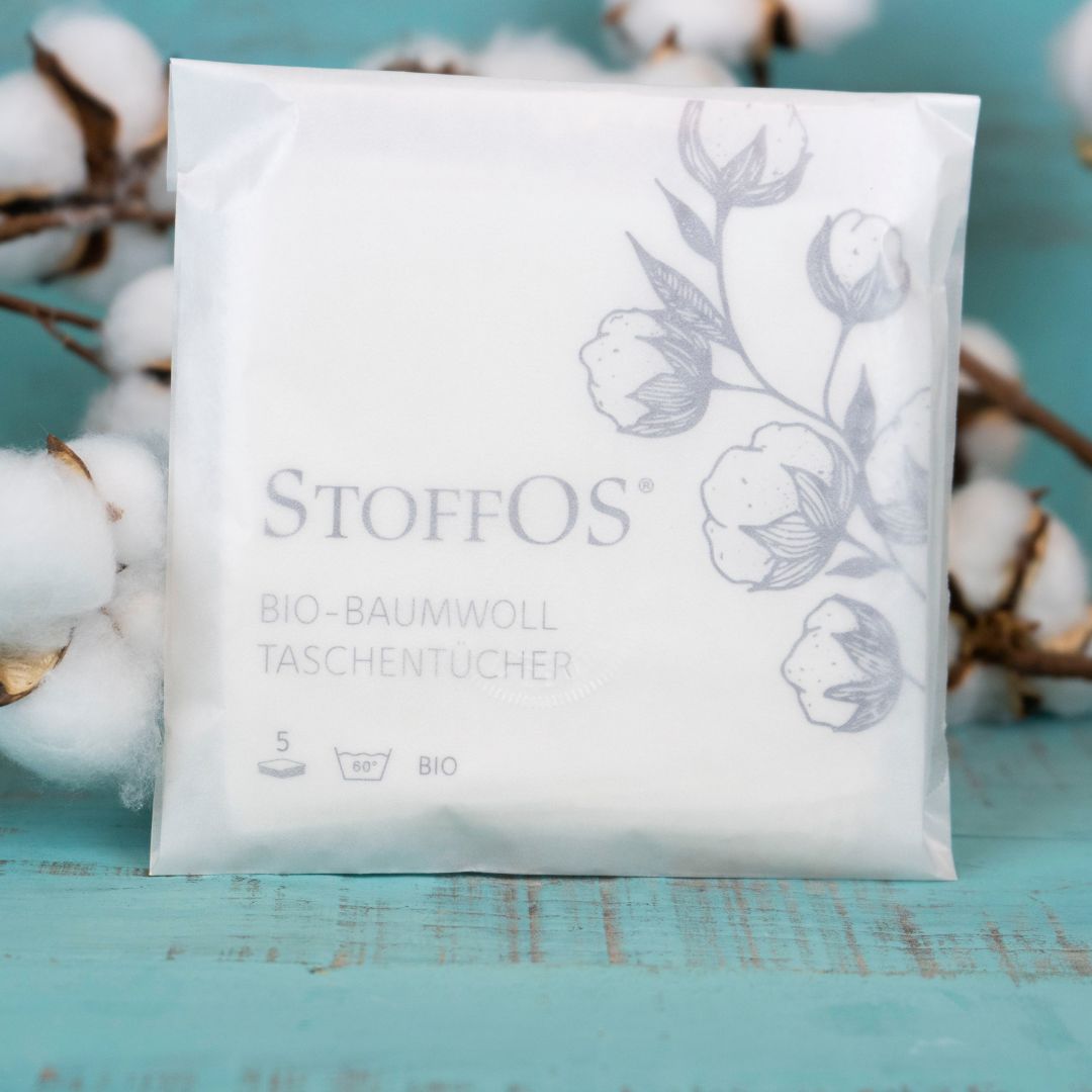 Stofftaschentücher, weiß, 5er Pack GOTS zertifizierten Bio-Baumwolljersey in einer halbtransparenten Pergamintüte. Unser zweites Verpackungsdesign: Das Verpackungsdesign ist etwas verspielter. Es zeigt auf dem rechten Drittel der Tüte einen gezeichneten Baumwollzweig mit sechs erntereifen Kapseln. Es wird deshalb gern statt Blumen verschenkt. Links davon das Logo StoffOS, mit Großbuchstaben OS am Ende.