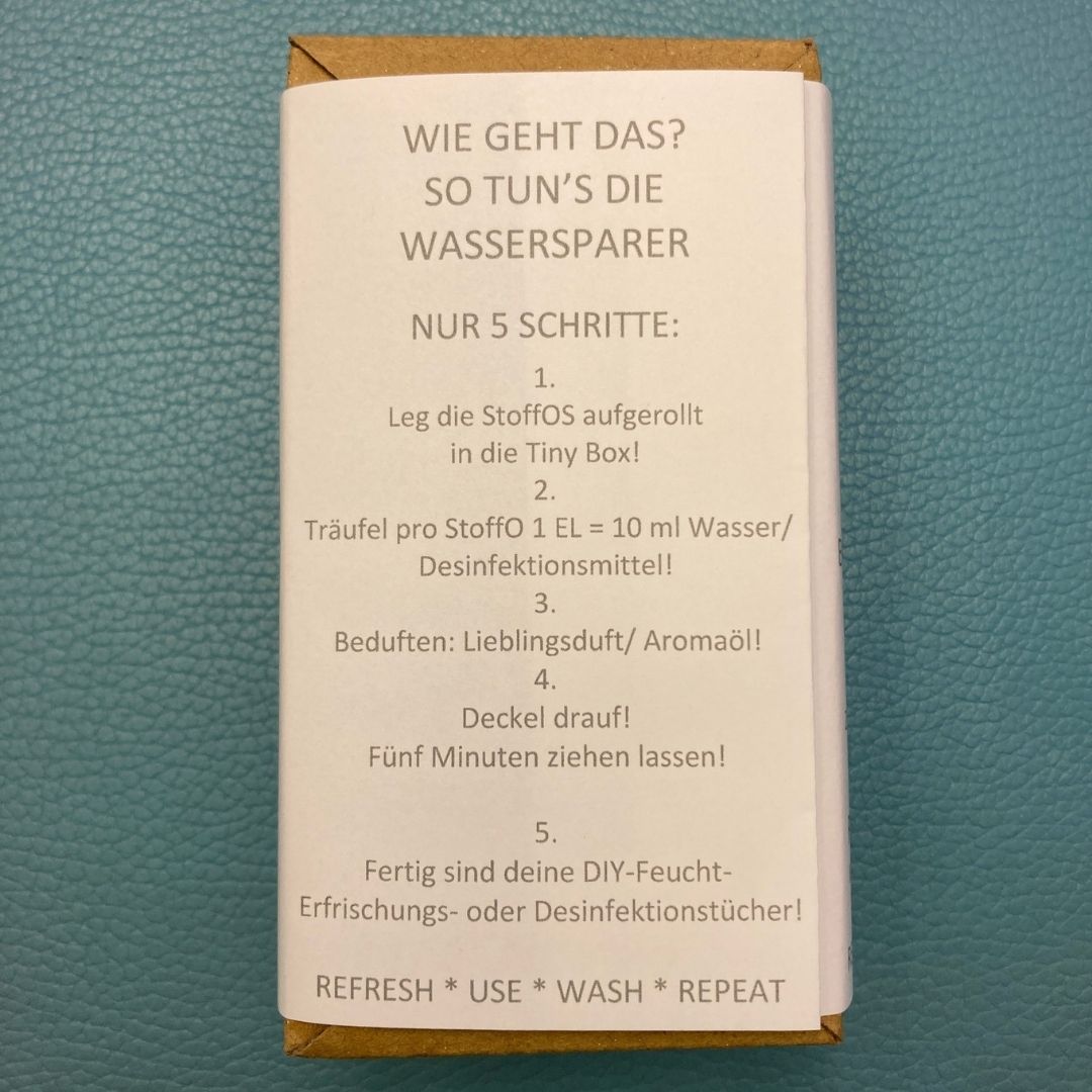 Rückseite der Banderole. Hier wird beschrieben, wie man in 5 Schritte aus den zusammenrrollten StoffOS in der Box mit jeweils einem Esslöffle Leitungswasser pro Röllchen Feuchttücher oder mit dem Lieblingsduft versehen, Erfrischungstücher herstellt. Statt Wasser Desinfektionsmittel verwenden, dann Desinfektionstücher.