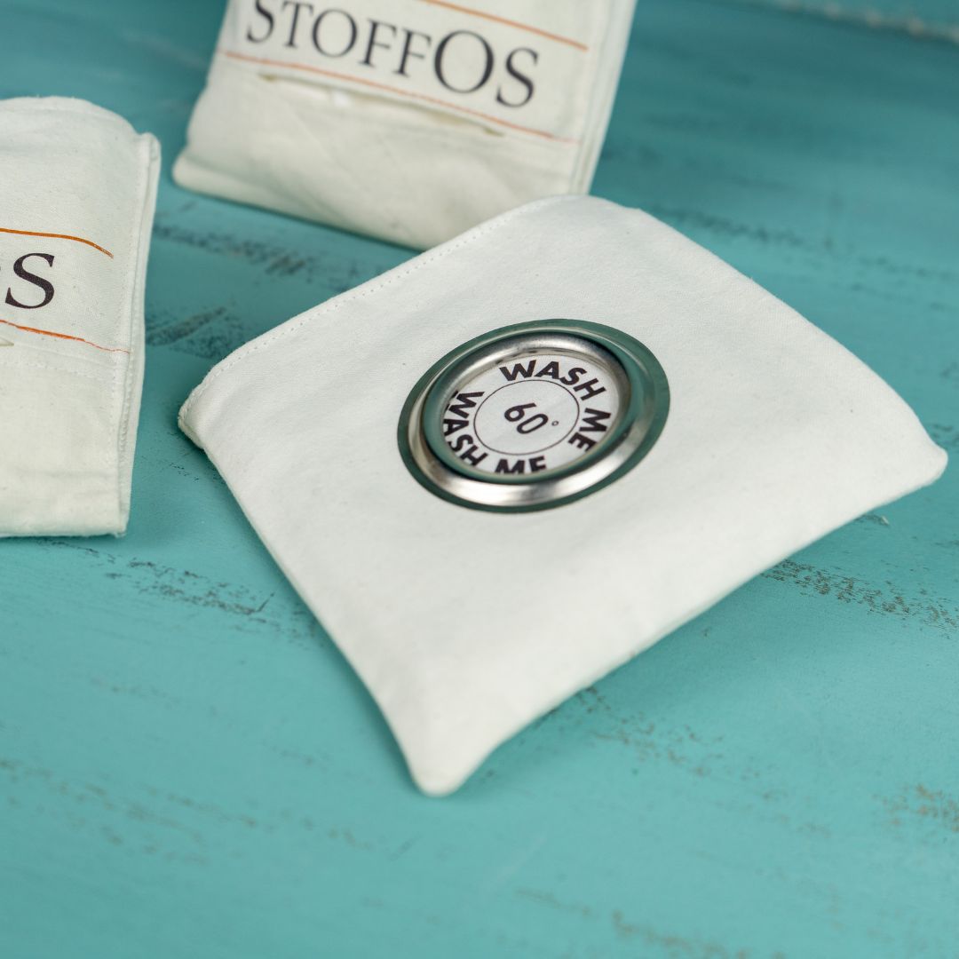 Auf der Rückseite der To Go Tasche mit vier Stofftaschentüchern befindet sich das dritte Fach. In der Mitte ist eine Edelstahlöse mit 6 cm Durchmesser eingearbeitet. Durch diese Öse sollen die vier benutzen StoffOS gesteckt werden. In der Mitte der Öse sieht man auf den innenliegenden Stoff, darauf ist in grau gedruckt: Wash me und 60° C.