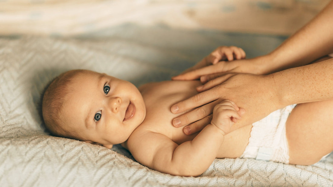 Ein ca. 3 Monate altes Baby nur mit einer Windel bekleidet liegt auf einer weichen UNterlage. Es lächelt und wird von zwei Frauenhänden am Bauch gespreichelt.