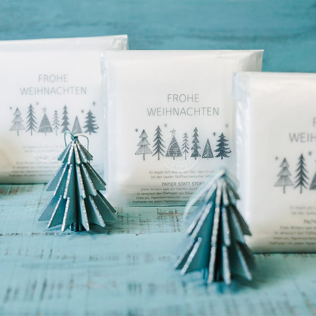 Die Pergamintüten stehen versetzt hintereinander. Dekoriert mit Mini Tannen aus Holz in matt türkis. Unser viertes Verpackungsdesign. Weihnachten. Fünf Stofftaschentücher aus weißem GOTS zertifizierten Bio-Baumwolljersey in einer halbtransparenten Pergamintüte. Über der Reihe von sieben unterschiedlich stilisierten Tannenbäumen in der Mitte der Verpackung steht FROHE WEIHNACHTEN. Darunter beginnt die Weihnachtsgeschichte. Sie wird bei den nächsten Bildern erwähnt werden.