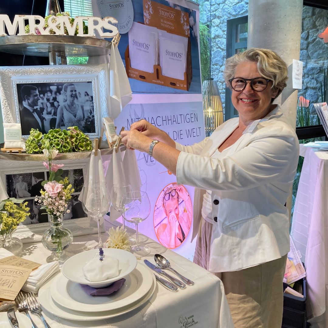 Messestand auf einer Hochzeitsmesse. Im Vordergrund ist ein Gedeck für ein mehrgängiges Menü ausgestellt. Im Teller liegt die StoffOS Stoffserviette, unser Nasenschmeichler, mit einer Lavendelblüte im Knopfloch der Serviette. Rechts daneben steht Silke Kretzing, die Gründerin von StoffOS. Sie dekoriert StoffOS Taschentücher am Rand einer Etagere.