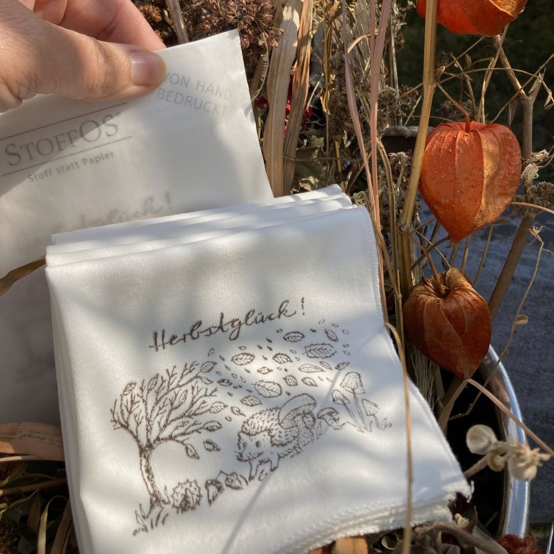 Stofftaschentücher HANDBEDRUCKT Fünf ausgepackte StoffOS mit dem Herbstglück Motiv bedruckt in kieselgrau liegen neben einer Pergamintüte mit Herbstglück-StoffOS in einem Herbsttrockenblumenstrauß in beige, braun und orange. Das Bild hat Nele Jamin von mach-werk aus der Igelperspektive gezeichnet. Links biegt sich ein Baum im Sturm, Blätter wehen durch die Luft. Am Boden die üblichen Herbstgefährten: Kastanien, Eicheln, Pilze und ein Igel.