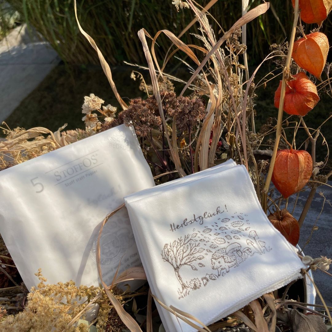 Stofftaschentücher HANDBEDRUCKT Fünf ausgepackte StoffOS mit dem Herbstglück Motiv bedruckt in kieselgrau liegen neben einer Pergamintüte mit Herbstglück-StoffOS in einem Herbsttrockenblumenstrauß in beige, braun und orange. Das Bild hat Nele Jamin von mach-werk aus der Igelperspektive gezeichnet. Links biegt sich ein Baum im Sturm, Blätter wehen durch die Luft. Am Boden die üblichen Herbstgefährten: Kastanien, Eicheln, Pilze und ein Igel.