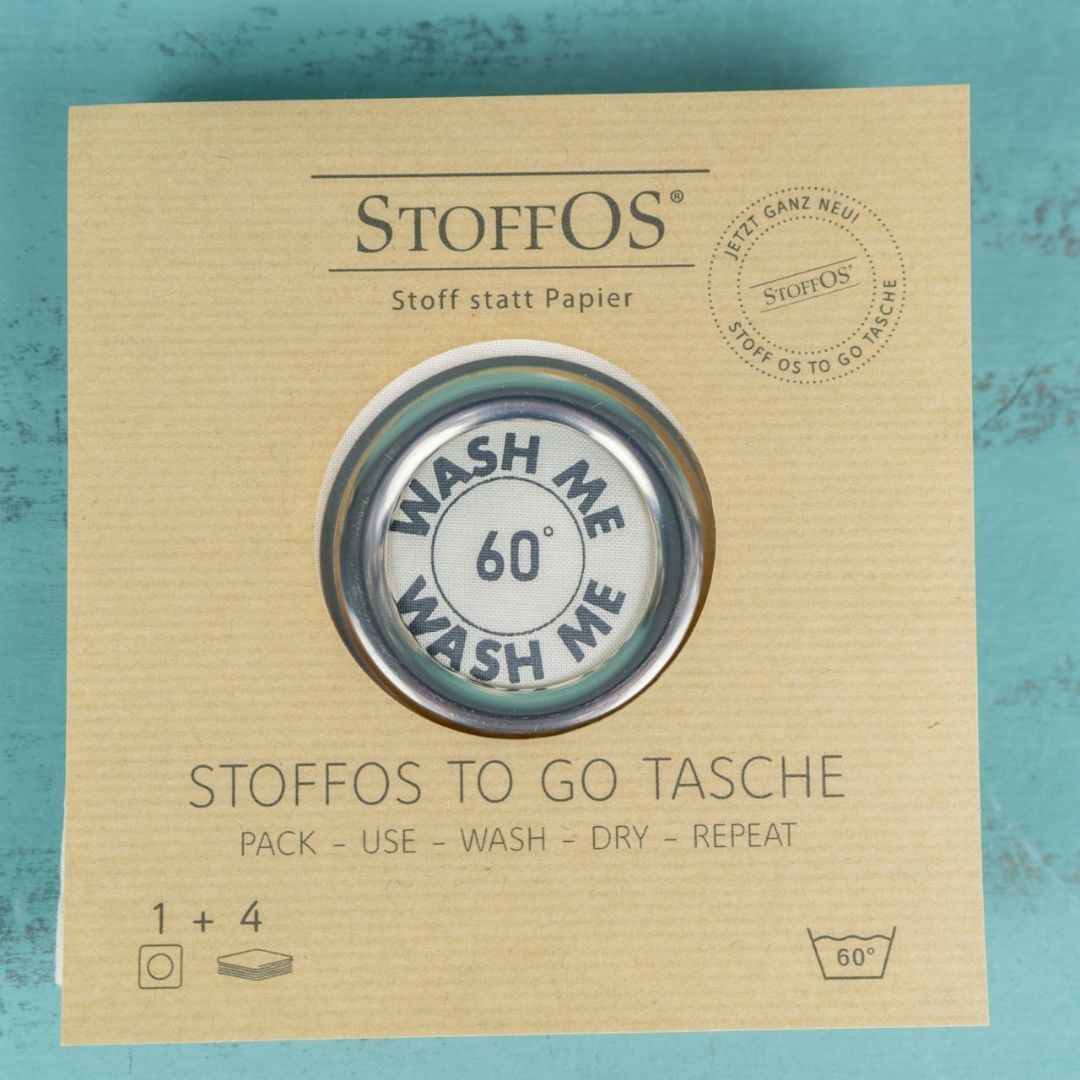 Die Stofftaschentücher Tasche liegt unter dem packpapierfarbenem Einlegerblatt mit dem Produktnamen: StoffOS To Go Tasche unten, StoffOS Logo oben und dem auf einem türkisfarbenen Untergrund. In der Mitte ist durch eine runde Ausstanzung der Blick auf die 6 cm große Edelstahlöse frei.