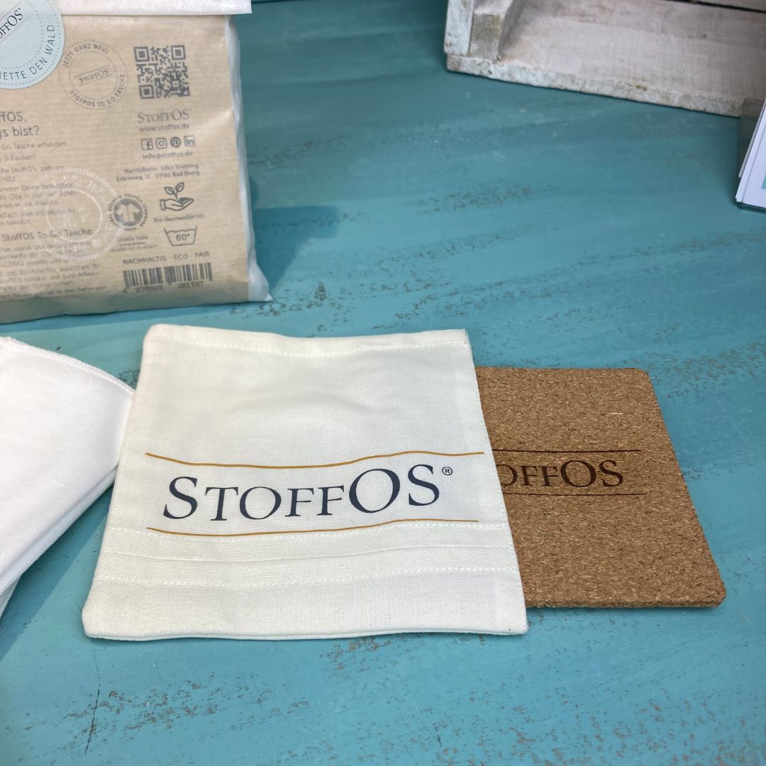Aus der Tasche, die liegend mit dem StoffOS Logo zu sehen ist, wurde die Korkplatte soweit herausgezogen, dass das Branding auf der Korkplatte zu sehen ist. Darauf ist gelasert: StoffOS Das entspricht dem Aufdruck auf der Frontseite  der Tasche.